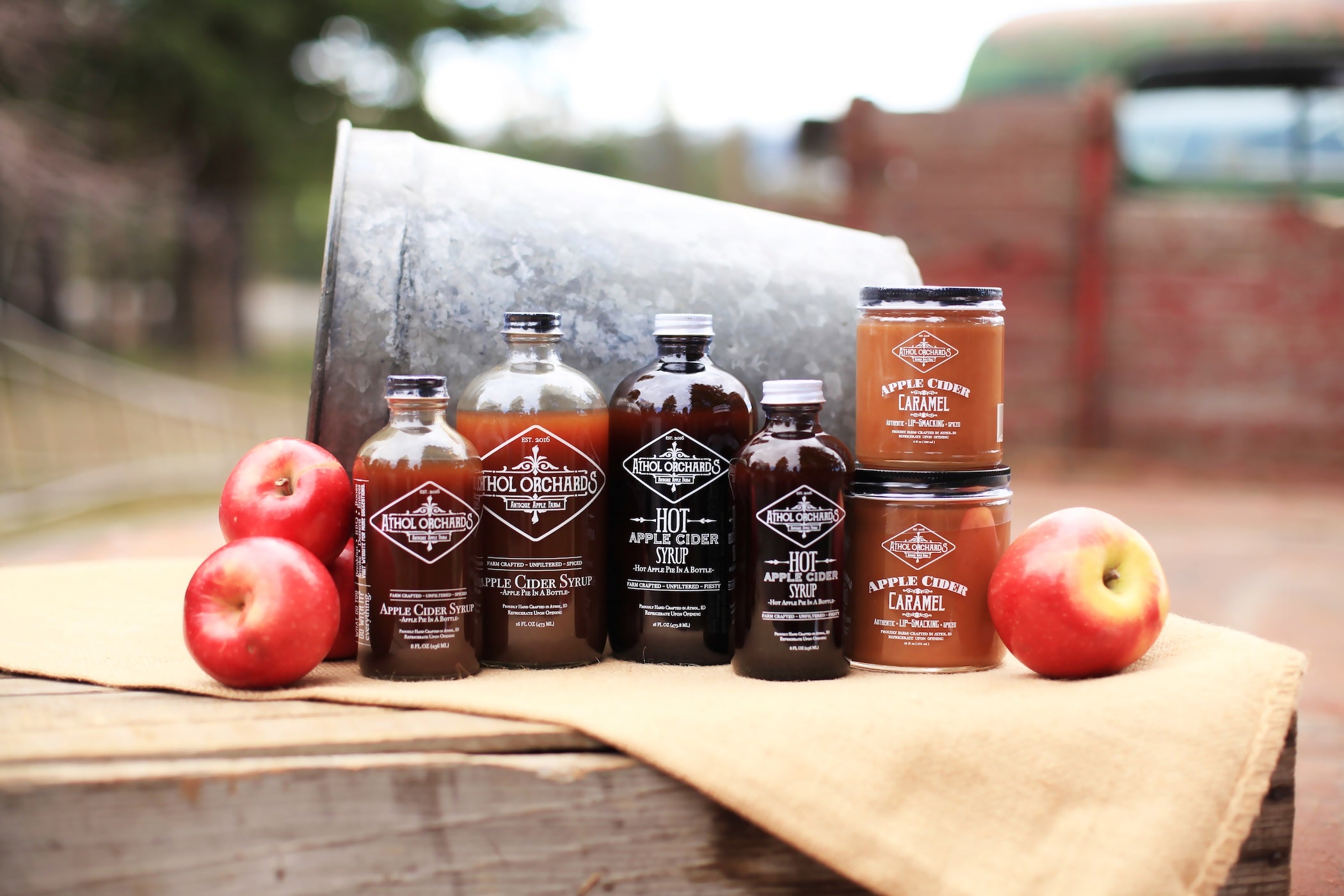 Collection of Apple Cider syrup products with apples on a wooden surface outdoors.